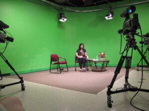 Woman seated in a green screen studio with cameras and lights around.