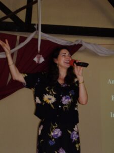 Woman singing into a microphone on stage with decorative backdrop.