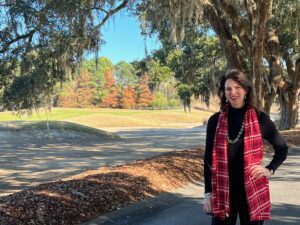 Woman in a red plaid vest standing on a sunny path with trees in fall colors.